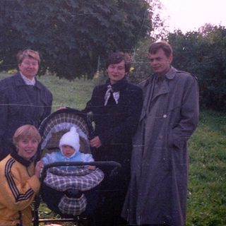 Family stands near a stroller in a grassy, wooded area.