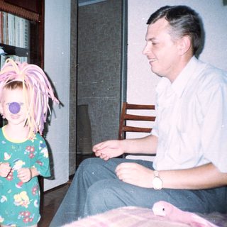 Child with a wig stands near a seated man.