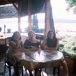 Three girls sit at a table beneath a shaded porch.