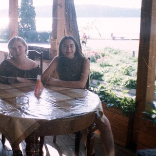 Three girls sit at a table beneath a shaded porch.