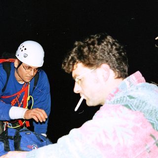 Two men, one climbing, the other smoking, in darkness.