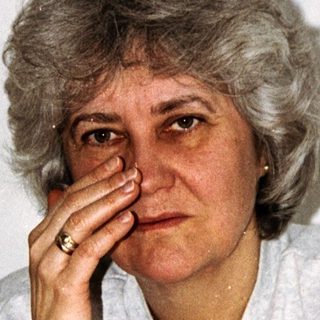 Here's a brief summary of the image: Older woman with short hair, touching her eye.