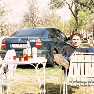 Man seated outdoors, next to a car and trees.