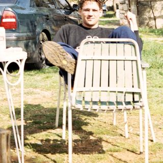 Man seated outdoors, next to a car and trees.