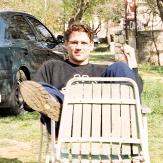 Man seated outdoors, next to a car and trees.