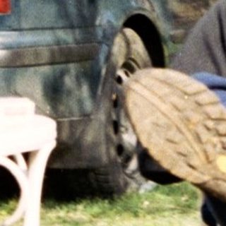 Man seated outdoors, next to a car and trees.