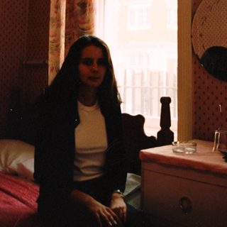 Woman sits near a window in a bedroom.