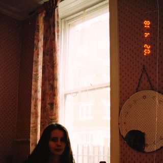 Woman sits near a window in a bedroom.