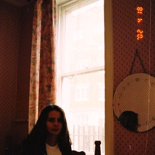 Woman sits near a window in a bedroom.