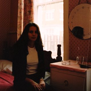 Woman sits near a window in a bedroom.