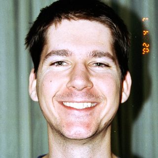 Here's a brief summary: Man smiling for a portrait, wearing a blue t-shirt.