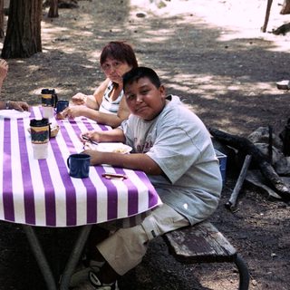 Family sits at a picnic table in a wooded area.