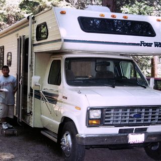 A boy stands near a vintage camper van in a forest.