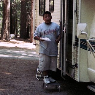 A boy stands near a vintage camper van in a forest.