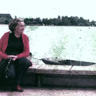 Woman sits by a lake, near boats and trees.