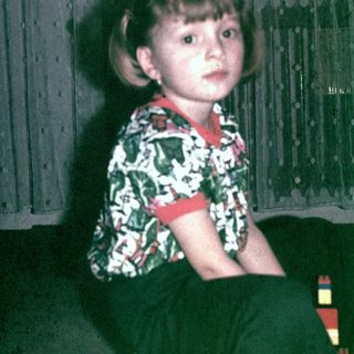 Girl wearing floral shirt, seated near a curtain.