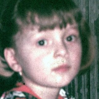 Girl wearing floral shirt, seated near a curtain.