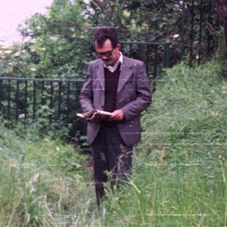 Man reading in tall grass with a lake view.