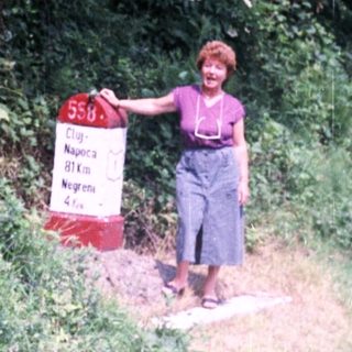 Woman stands near a road sign in a wooded area.