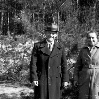 Couple stands outdoors in coats, posing in a wooded area.