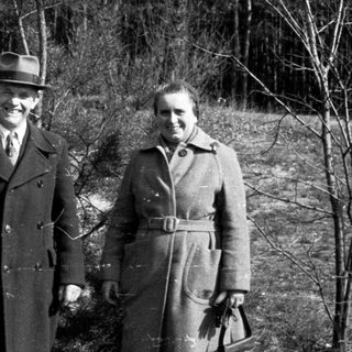 Couple stands outdoors in coats, posing in a wooded area.