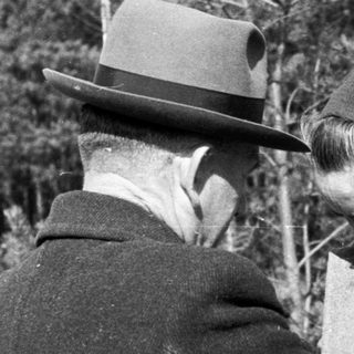 Man and woman in coats stand outdoors in a wooded area.