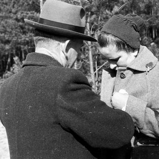 Man and woman in coats stand outdoors in a wooded area.