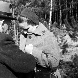 Man and woman in coats stand outdoors in a wooded area.