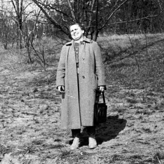 Woman in coat stands in a barren, wooded landscape.