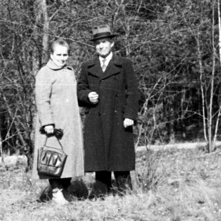 Here's a brief summary of the image: Couple stands in a grassy, wooded area.