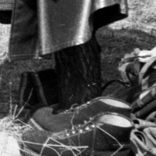 Man sits near clothing and luggage in a wooded area.