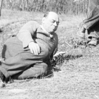 Man sits near clothing and luggage in a wooded area.