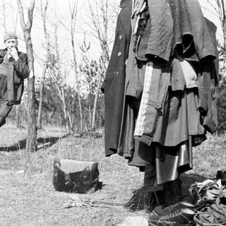 Man sits near clothing and luggage in a wooded area.