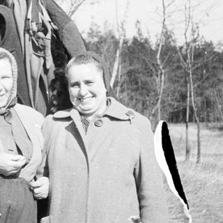 Two women in coats standing outdoors in a wooded area.