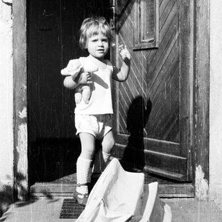 Here's a brief summary of the image: Baby stands outside a door, holding a toy.