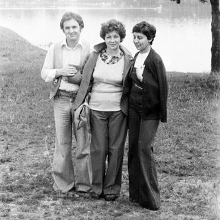 Three people stand near a body of water, in a field.