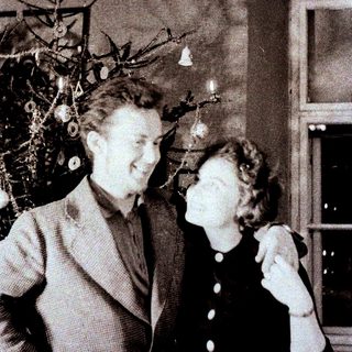 Couple stands near decorated Christmas tree in vintage setting.