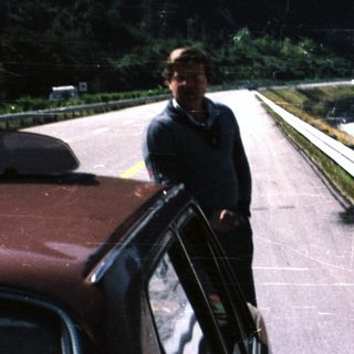Man stands next to a car, overlooking a river and mountains.
