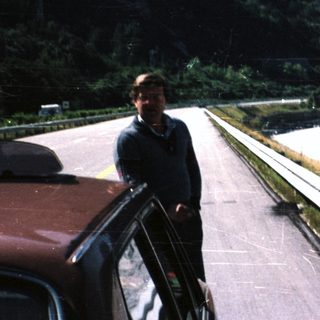 Man stands next to a car, overlooking a river and mountains.
