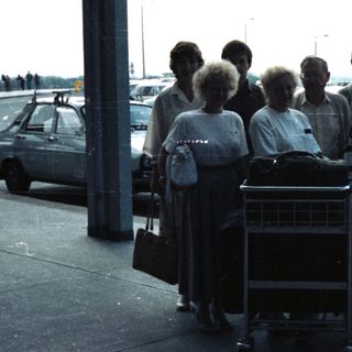 Group of people and luggage stand under an airport canopy.