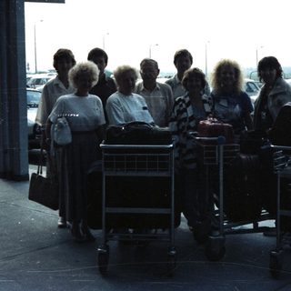 Group of people and luggage stand under an airport canopy.