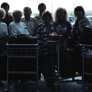 Group of people and luggage stand under an airport canopy.
