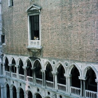Here's a brief summary of the image: Arched facade of a historic building with people on a balcony.