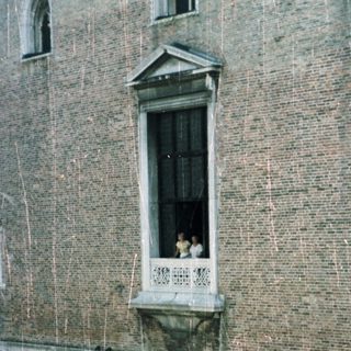 Here's a brief summary of the image: Arched facade of a historic building with people on a balcony.