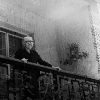 Elderly man stands on a balcony, looking away from camera.
