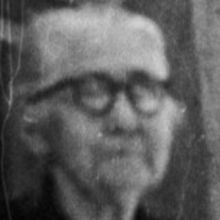 Elderly man stands on a balcony, looking away from camera.