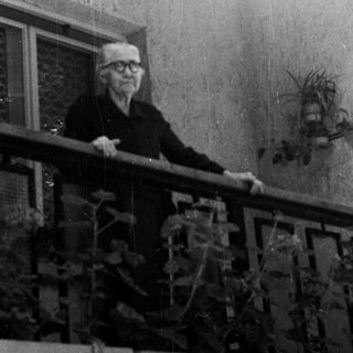 Elderly man stands on a balcony, looking away from camera.