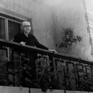 Elderly man stands on a balcony, looking away from camera.