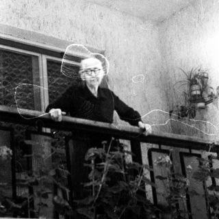 Here's a brief summary of the image: Older woman standing on a balcony with plants.