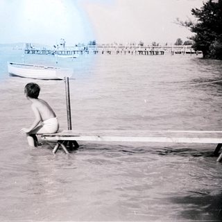 Boy sits on dock in water, boats and shoreline visible.